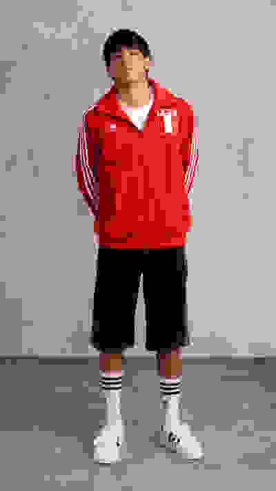 A young person wearing a red sports jacket with the Peruvian flag patch, standing in front of a plain gray background.