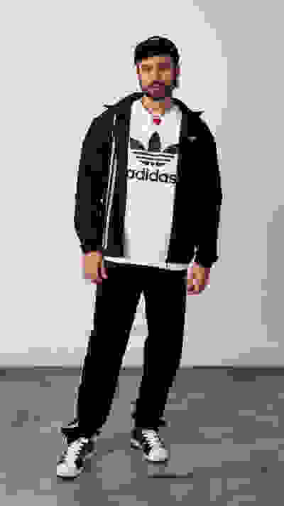 A man wearing an Adidas t-shirt, black jacket, and pants, standing in a plain room with a concrete floor.