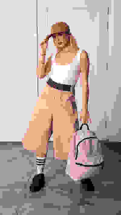 A young woman wearing a white tank top, beige wide-leg pants, and a baseball cap stands in a room with a plain white wall in the background, holding a backpack.
