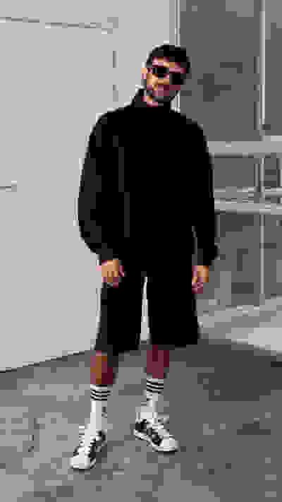 A person wearing a black sweater, shorts, and white sneakers stands in a room with a window in the background.