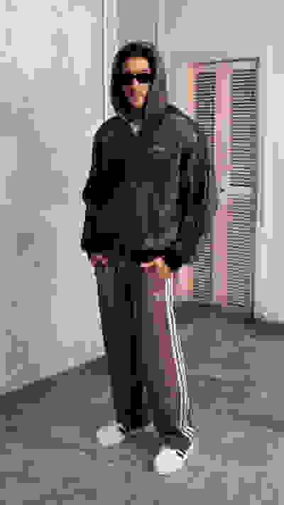 A person wearing a hooded camouflage jacket and sweatpants stands in a room with a concrete floor and a set of louvered doors in the background.