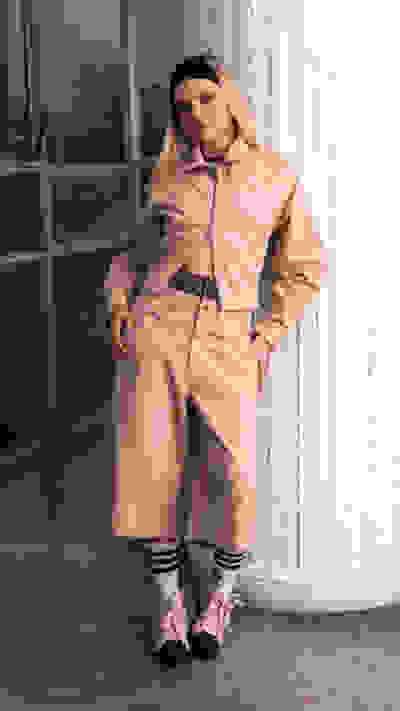 A person wearing a beige hooded jacket and pants stands in front of a window, with a brick wall visible in the background.