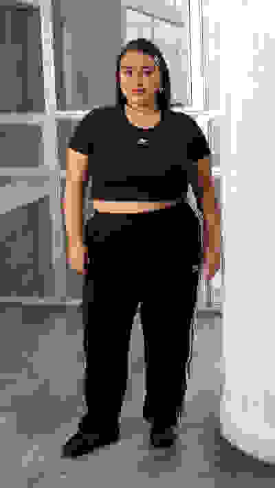 The image shows a person wearing a black crop top and black sweatpants, standing in a room with large windows and a concrete floor.