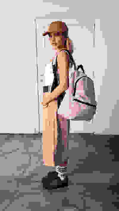 A young woman wearing a beige outfit, including a backpack, stands in a room with a plain white wall in the background.