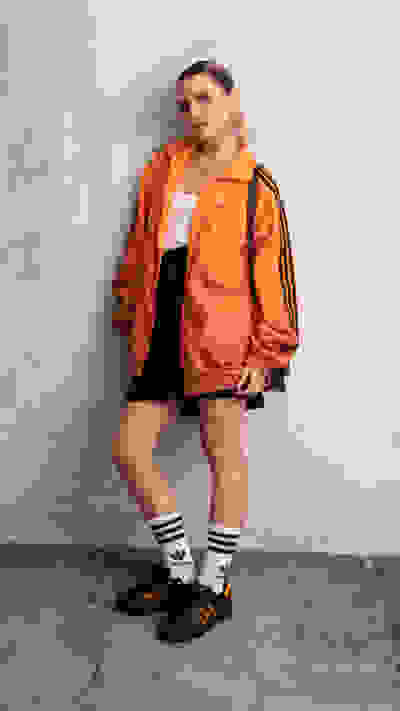 A person wearing an orange jacket, a white top, and black shorts stands in front of a plain gray wall.
