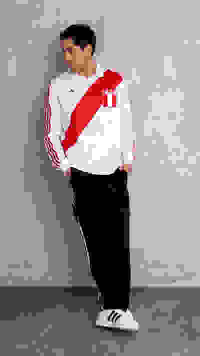 A young man wearing a white and red sports jersey stands against a plain gray wall, with black pants and white sneakers.