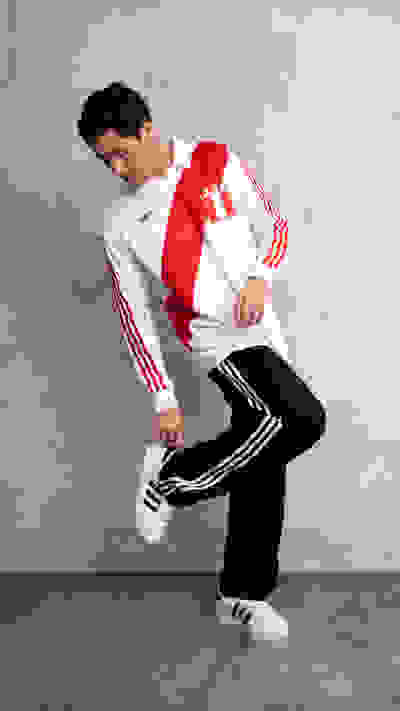 A young person wearing a white and red athletic jacket and black pants is standing in a plain, gray-toned background, appearing to be in a dynamic pose.