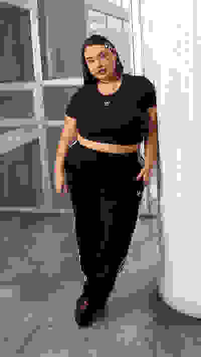 A woman wearing a black crop top and matching sweatpants stands in a room with concrete floors and large windows.