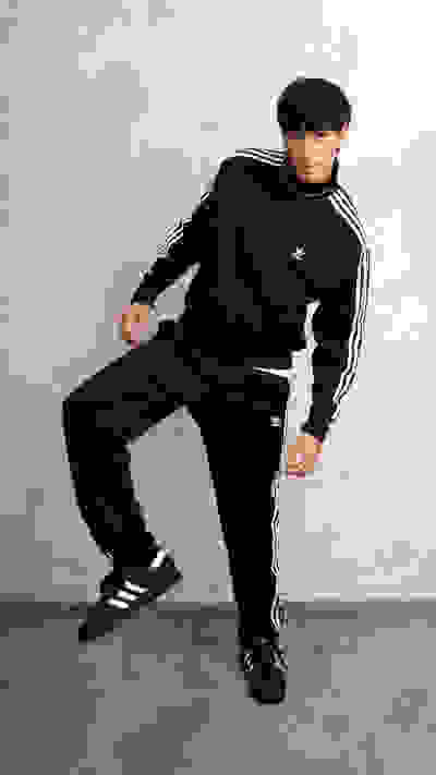 A young man wearing a black Adidas tracksuit stands against a plain, light-colored background, striking a casual pose.