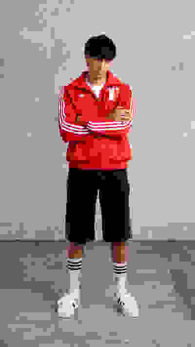 A young person wearing a red sports jacket and black shorts stands in front of a plain concrete wall.