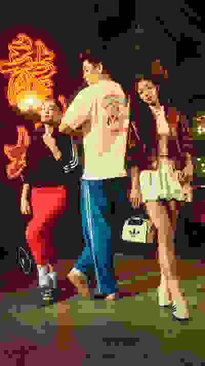 Three individuals, two women and one man, are standing in front of a neon sign in a dimly lit setting, dressed in colorful, stylized clothing that appears to be inspired by 1980s or 1990s fashion trends.