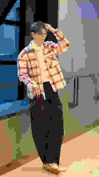 A young person wearing a plaid shirt, brown pants, and light-colored shoes stands in a room with glass walls and a tiled floor.