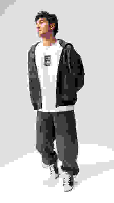 A young man wearing a white t-shirt, a camouflage jacket, and cargo pants stands against a plain white background.