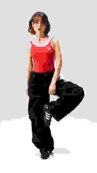 A young woman with curly hair wearing a red tank top and black pants stands against a plain white background, her expression conveying a sense of confidence and energy.