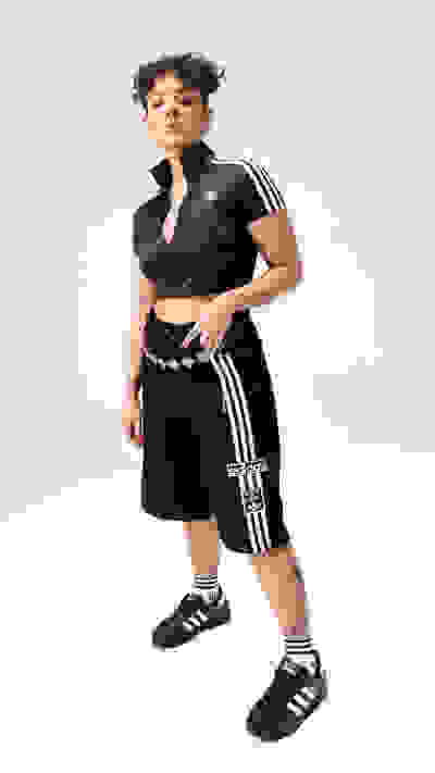 A young person with curly hair wearing a black athletic outfit and Adidas sneakers stands against a plain white background.