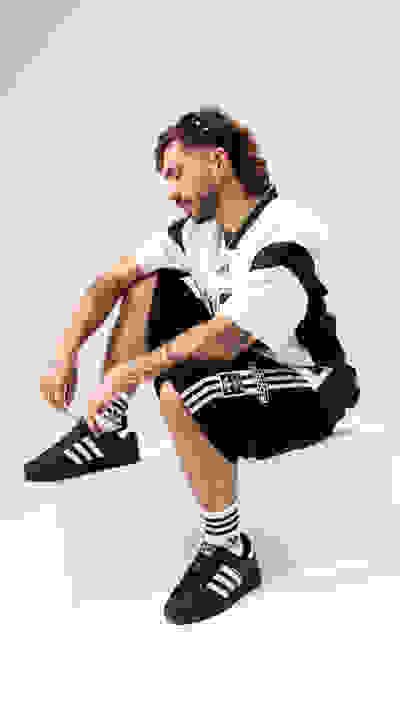 A man with long hair and tattoos sits on a white surface, wearing a black and white athletic jacket, black shorts, and black and white sneakers.