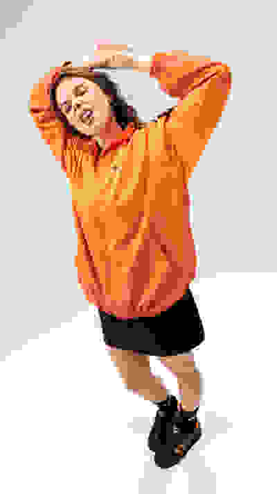 A young woman wearing an orange jacket stands with her arms raised, her expression conveying a sense of energy and enthusiasm against a plain background.