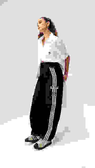 A person wearing a white blouse and black pants with stripes sits on a white platform against a plain background.