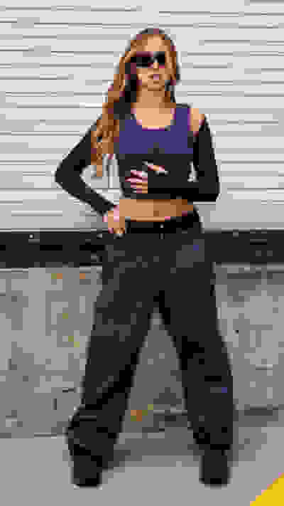 A young woman wearing a purple crop top and matching pants stands in front of a gray concrete wall, her long blonde hair and sunglasses adding to her stylish appearance.