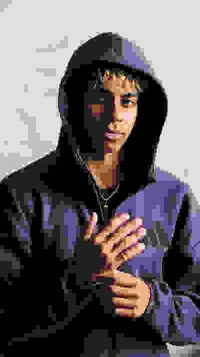 A young person wearing a purple hooded sweatshirt stands against a plain background, their hands clasped in front of them.