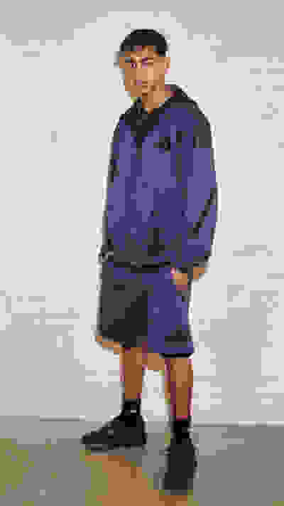 A young man wearing a purple tracksuit stands in front of a plain white wall.