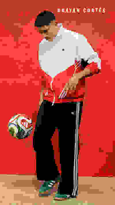 A young man in a white and red athletic jacket stands in front of a bright red background, holding a soccer ball.