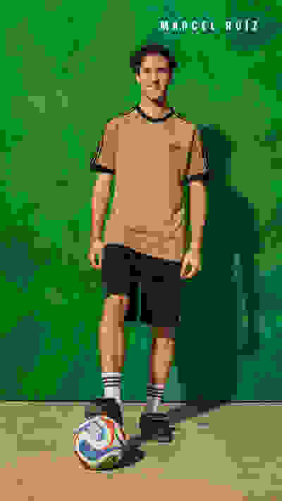 A young man wearing a tan t-shirt and black shorts stands in front of a vibrant green background, holding a soccer ball.