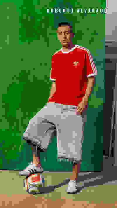 A man in a red Adidas shirt and gray shorts stands next to a soccer ball on a green background.