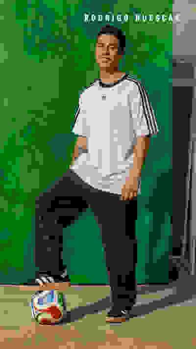 The image shows a young man wearing a white t-shirt and black pants, sitting on the floor next to a soccer ball in front of a green background with the name "RODRIGO HUESCAS" displayed.