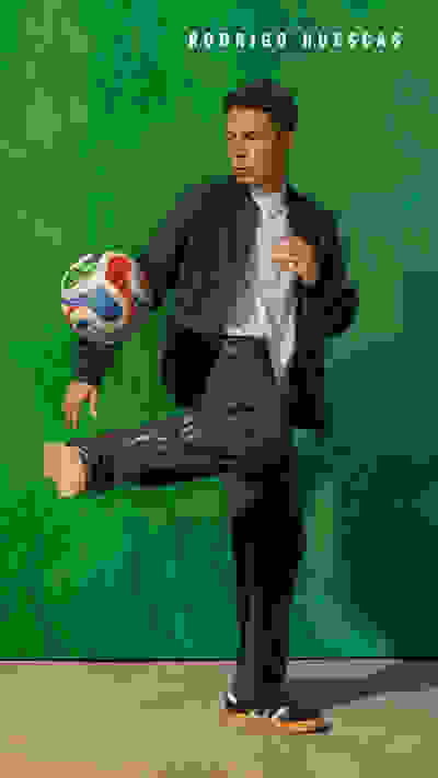 A man in a suit is holding a soccer ball and posing in front of a vibrant green background.