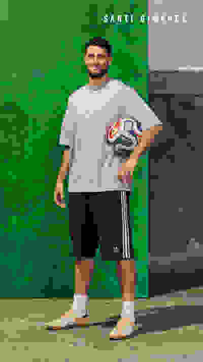 A young man in a white t-shirt and black shorts stands in front of a green background, holding a soccer ball.