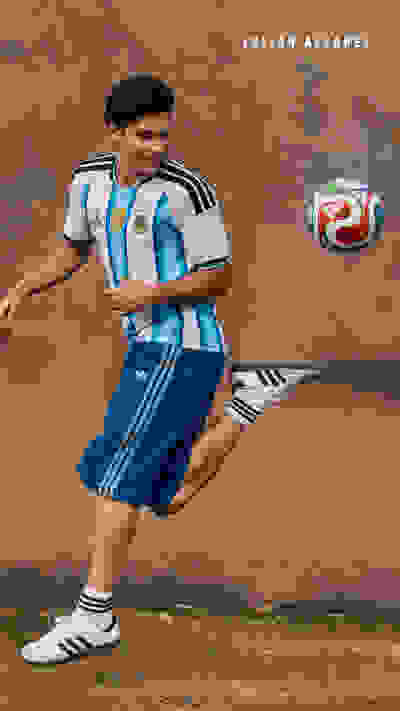 A young soccer player in an Argentina national team jersey stands in a dusty, outdoor setting, holding a soccer ball and appearing ready to take a shot.