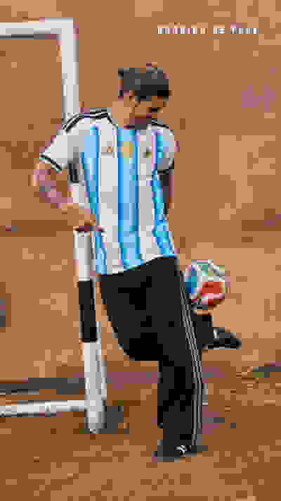 A man wearing an Argentina national team jersey and black pants stands in front of a textured wall, holding a soccer ball.