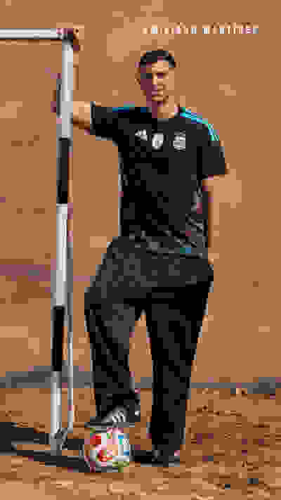 A man in a black sports jersey and pants stands in a desert-like setting, holding a baseball bat and standing next to a baseball.