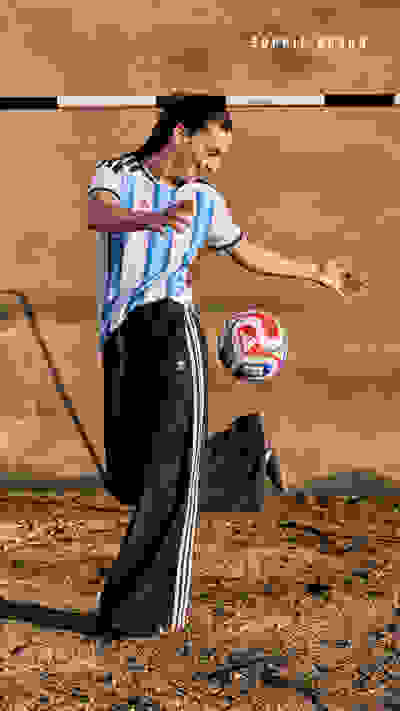 A person in a soccer jersey and black pants is kicking a soccer ball on a dirt field, with a backdrop of a sandy, arid landscape.