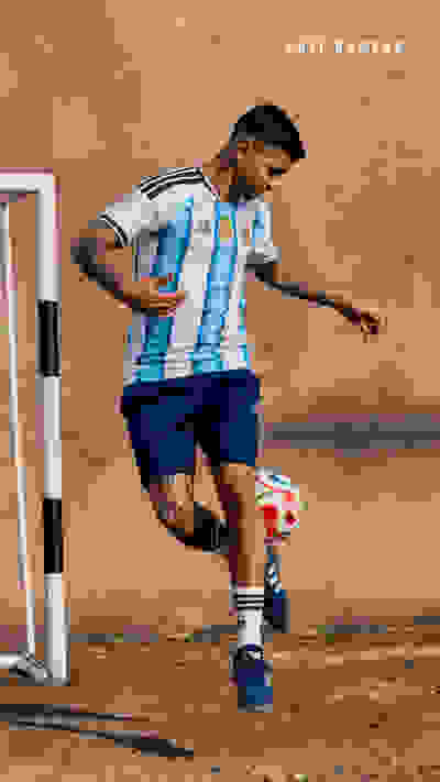 A soccer player in an Argentina jersey is dribbling a ball on a dirt field with a plain background.