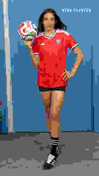 A woman in a red soccer jersey and shorts stands in front of a blue wall, holding a soccer ball.