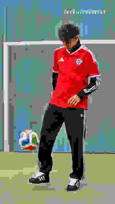 A person wearing a red soccer jersey and black pants stands in front of a blue metal wall, holding a soccer ball.
