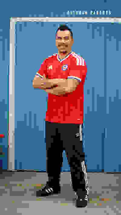 A smiling man wearing a red soccer jersey and black pants stands in front of a blue corrugated metal wall.