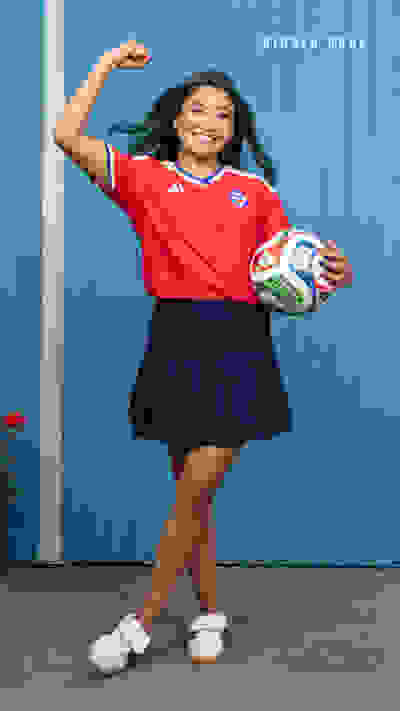A young woman in a red sports jersey and navy blue skirt stands in front of a blue wall, holding a soccer ball and smiling.