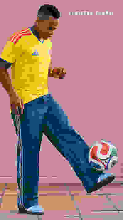 A young man in a yellow sports jersey is standing in front of a pink wall, casually controlling a soccer ball with his feet.