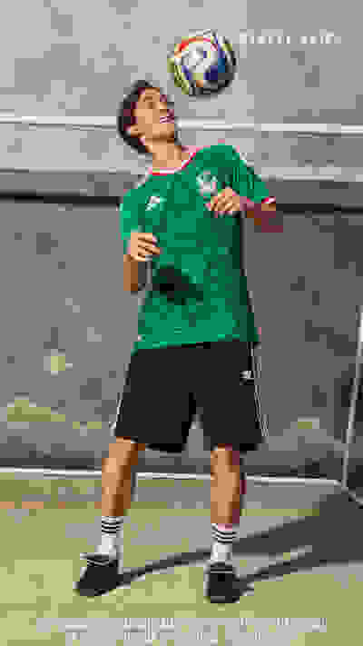 A young man in a green jersey with a soccer ball is standing in front of a mural depicting a scenic landscape.