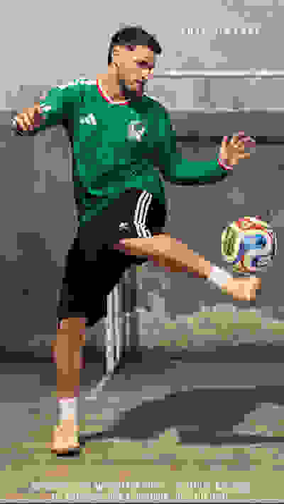 A soccer player in a green jersey is mid-kick, with a soccer ball in the air, against a wall with the name "Santi Gimenez" written on it.