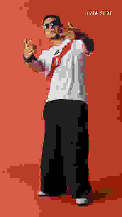 A person wearing a white t-shirt with a Peruvian flag, sunglasses, and loose black pants is gesturing with their hands against a bright red background.