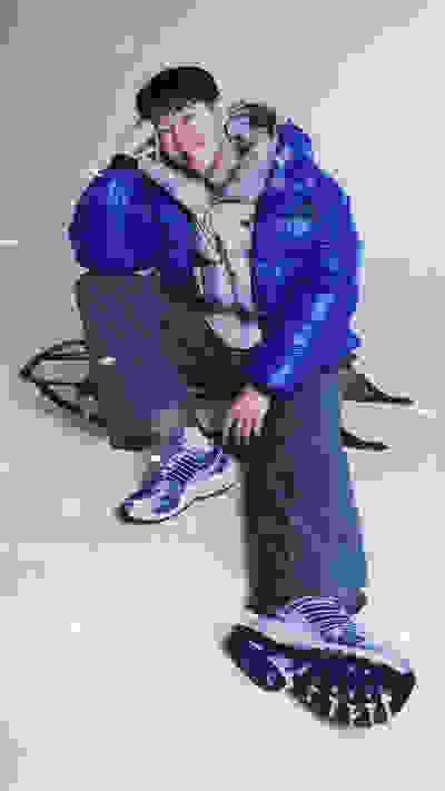 A young person wearing a vibrant blue puffy jacket and sneakers sits on a snowy surface, with a blurred background suggesting a wintry outdoor setting.