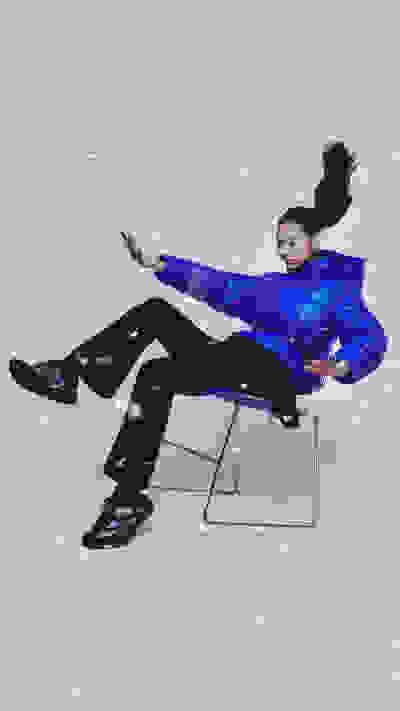 A person in a vibrant blue jacket is sitting on a chair, their arms outstretched, against a snowy background.