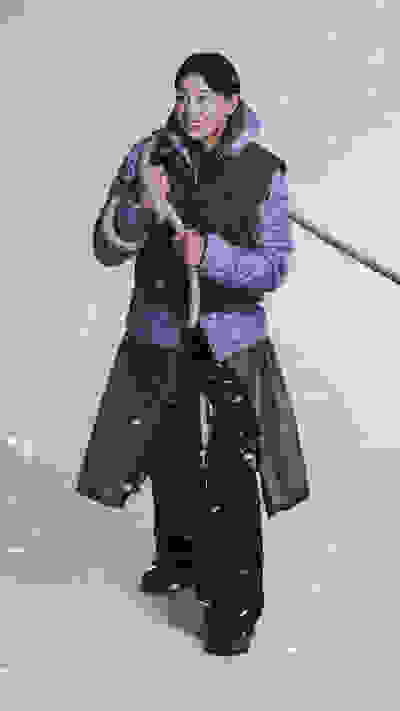 A person in warm winter clothing is standing in a snowy outdoor environment, holding a rope and appearing to be engaged in some kind of activity or task.