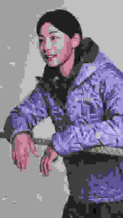 A person wearing a gray and olive green winter jacket stands in a snowy outdoor setting, with a rope visible in the foreground.