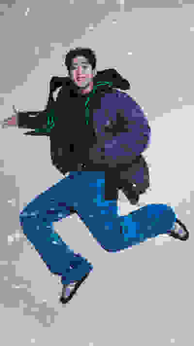 A person wearing a black jacket and blue pants is jumping in the air against a snowy background.