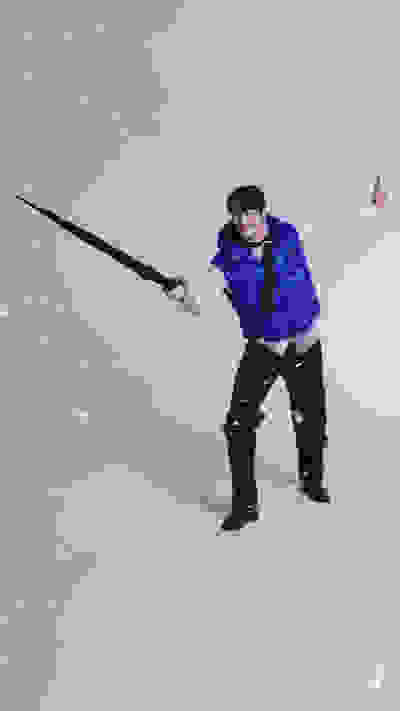 A person in a blue jacket and winter gear stands in a snowy landscape, holding a long pole and making a gesture with their free hand.